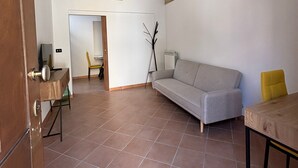 A spacious living room with a grey sofa, a wooden table, and a yellow chair. There is a door with a wooden frame and a radiator on the wall. - Casaggiù traditional house just steps from the towncenter & seaside destinations (San Vito dei Normanni)