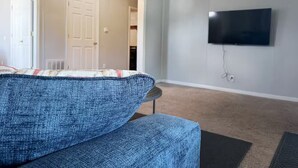 Een woonkamer met een blauwe bank, een aan de muur bevestigde televisie, en een deur naar een andere kamer.