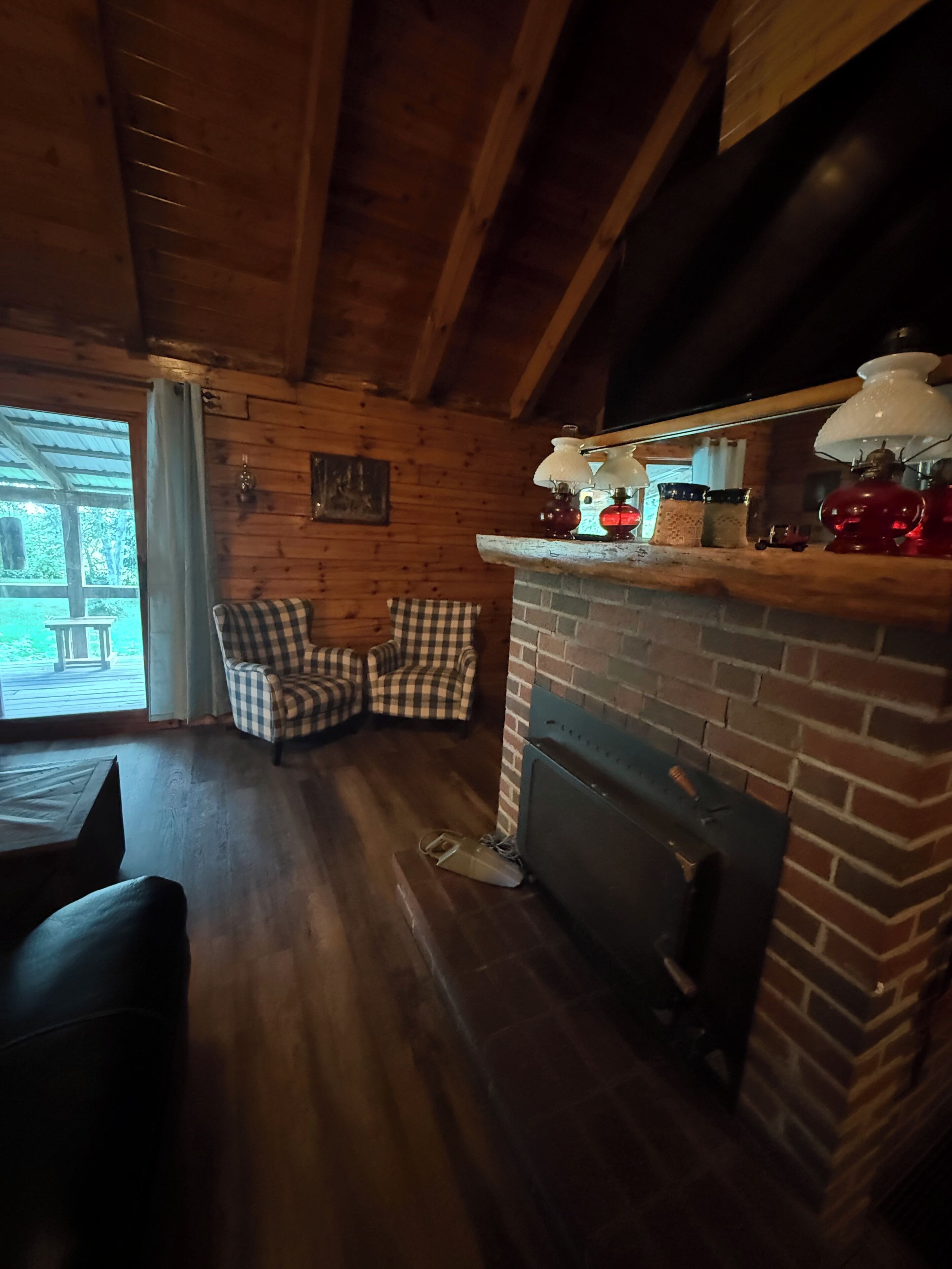 L'interno accogliente di una baita con soffitto in legno, una poltrona a quadri, un caminetto e una finestra vista su spazi verdi.