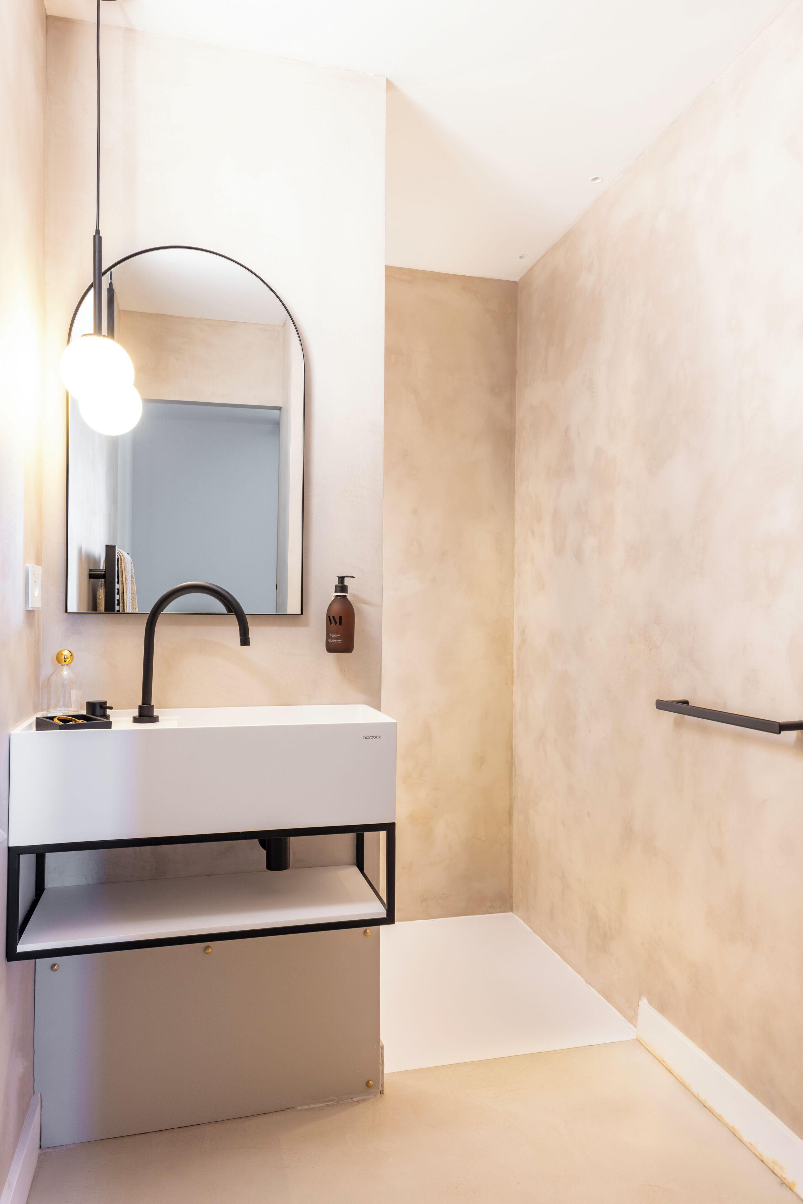 Un baño moderno con un lavamanos blanco, un grifo negro, un espejo redondo y una sola botella de champú.