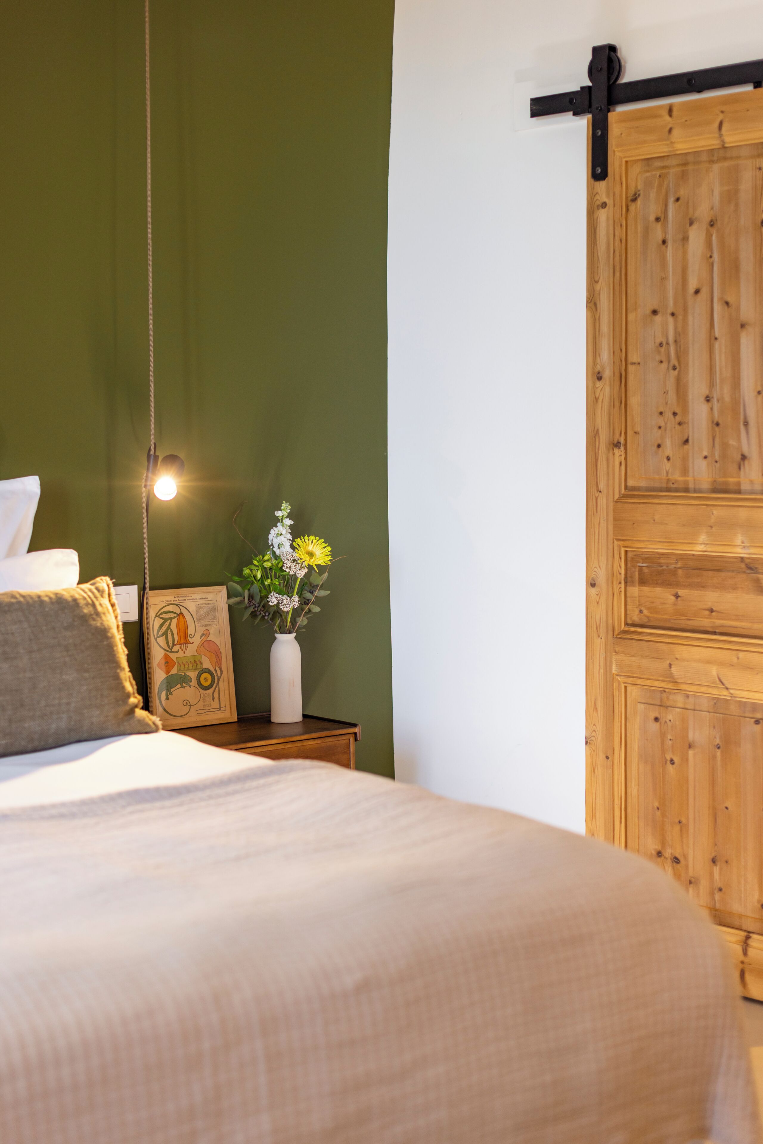A bedroom with a bed, a nightstand with a vase of flowers, a wooden door, and a wall-mounted light.