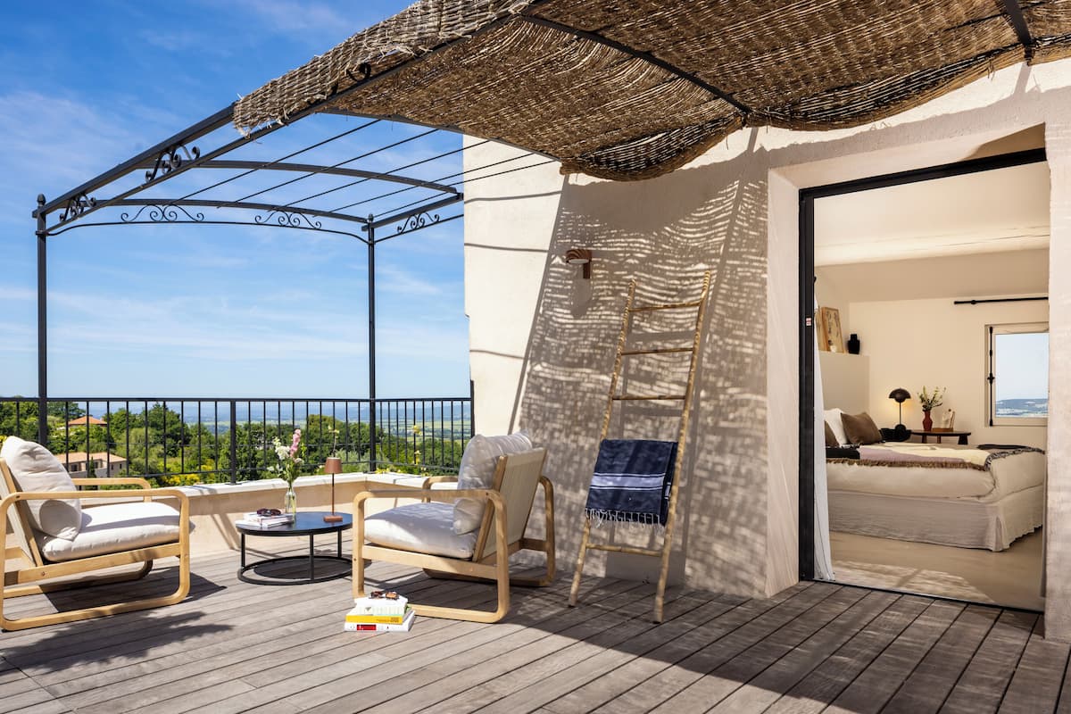Un balcón con piso de madera, un parasol de mimbre, dos sillones de descanso, una mesa redonda pequeña y vista al mar.