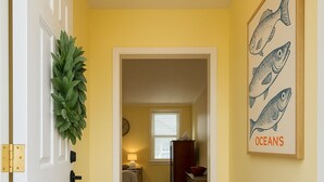 A hallway with a striped carpet, a door with a brass handle, a small stool, a wall-mounted plant, and a framed fish illustration. - Lake Breeze Cottage.   
One block from the lake, walking distance to downtown! (Sheboygan)