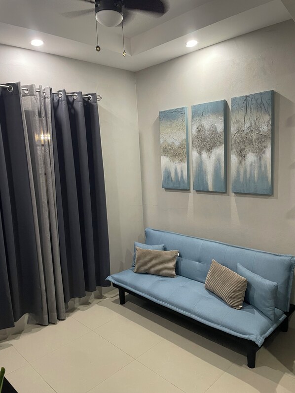 A room with a blue sofa, two paintings on the wall, a ceiling fan, and a curtain.