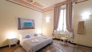 A hotel room with a bed, a chair, a window with curtains, a radiator, and a painting on the wall. - Genova Principe Suite (Genoa)