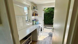A narrow kitchen with a washing machine, a countertop with various items, and a view of a garden. - Detached country house in Drenthe with large garden, terrace & beautiful views (Schoonloo)