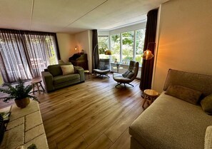 A living room with a sofa, armchairs, a piano, and a large window with curtains. - Detached country house in Drenthe with large garden, terrace & beautiful views (Schoonloo)