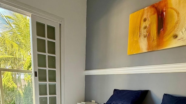 A neatly made bed with a folded towel, a nightstand, a painting on the wall, and a window with a view of greenery.