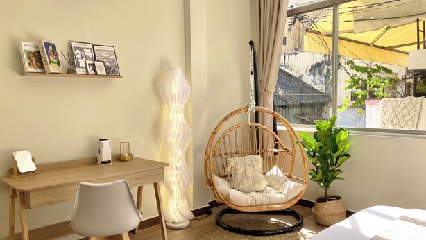 A room with a wooden desk, a white chair, a hanging chair with cushions, a floor lamp, a potted plant, and a bed with a patterned bedspread. - Romantic Studio with Sunlight in the City Center (Hồ Chí Minh)