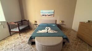 A bedroom with a bed, a crib, a round ottoman, a painting on the wall, and a dresser. - Villa Sole (Petrosino)