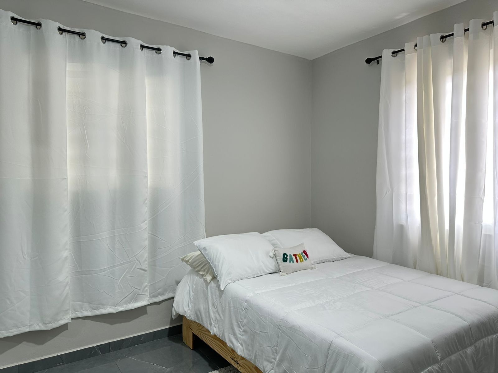 A bedroom with a bed, white curtains, and a pillow with the word "GATHER".