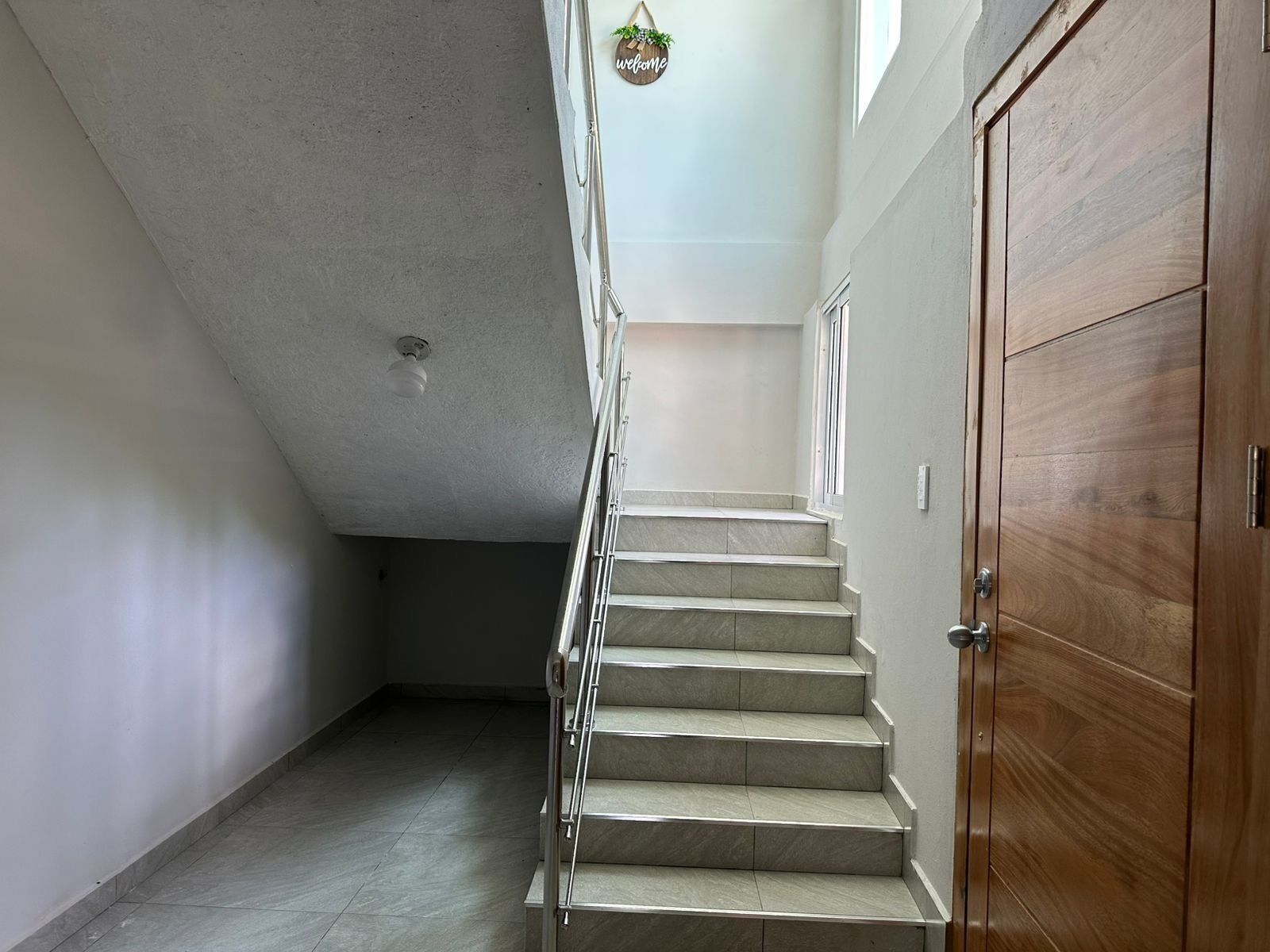 Una escalera con una puerta de madera a la derecha, una lámpara en el techo y un cartel que dice 'welcome'.