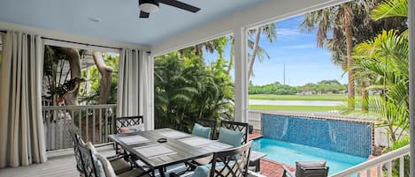 A covered outdoor area with a dining table and chairs, a ceiling fan, and a view of a swimming pool and greenery.