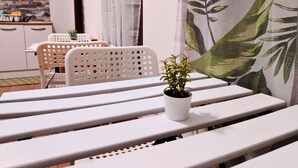 Et spiseområde med hvide bænke og stole, en lille potteplante på en bænk, og en væg med bladmotiv.