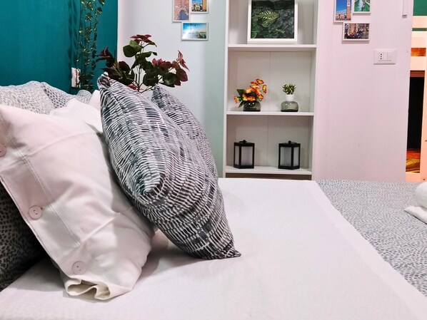 Ein ordentlich bezogenes Bett mit gemusterten Kissen, ein weißer Nachttisch mit Blumenvase sowie ein Regal mit gerahmten Bildern und kleinen Laternen.