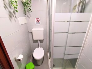 A small, white-tiled bathroom with a toilet, a green trash bin, and a no-smoking sign. - Charm Double Room Magnolia with private bathroom (Venezia)