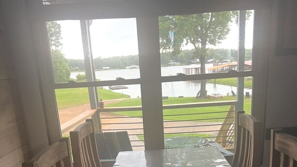 A dining table with chairs, a glass, and a pitcher, set against a view of a lake and grassy area. - Fisherman’s paradise waterfront. Boat ramp on property sleeps 6.      2-2.  (Many)
