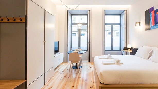A modern hotel room with a wooden floor, a bed with white linens, a desk with a chair, and a television. - Modern Retro Downtown by LovelyStay (Porto)