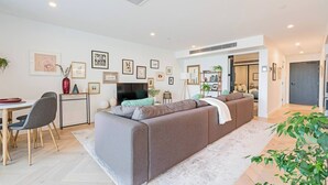 A modern living room with a grey sofa, a dining area with a table and chairs, and a hallway leading to a bedroom.