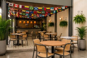 An outdoor dining area with round tables and wicker chairs, a bar with bottles, and potted plants. Flags are hung above the tables. - Capybara Hostel (Villavicencio)