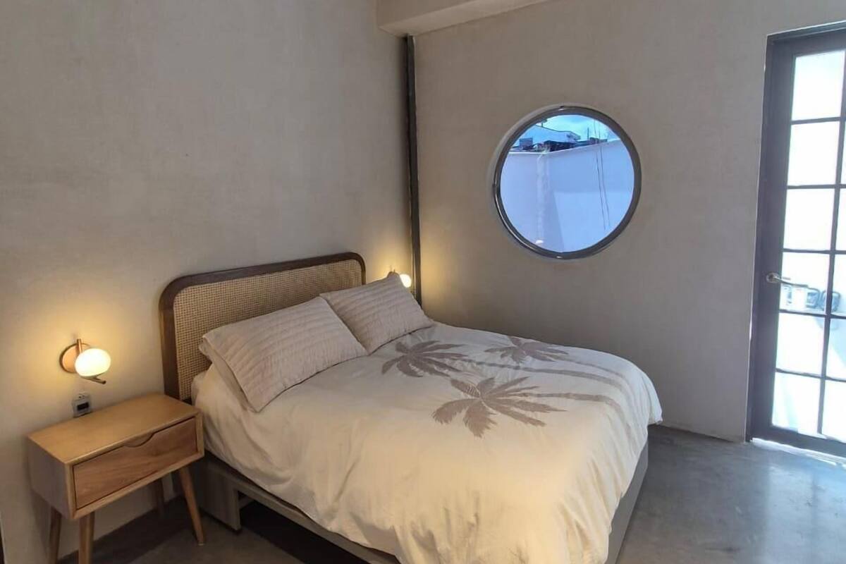 A bedroom with a bed, a nightstand, a round mirror, and a window.