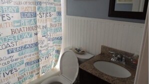 A bathroom with a shower curtain featuring nautical words, a white toilet, a granite sink, and framed pictures on the wall. - Oceanside, Duplex - 1st Flr Unit North Beach Haven, 20th St (Long Beach)