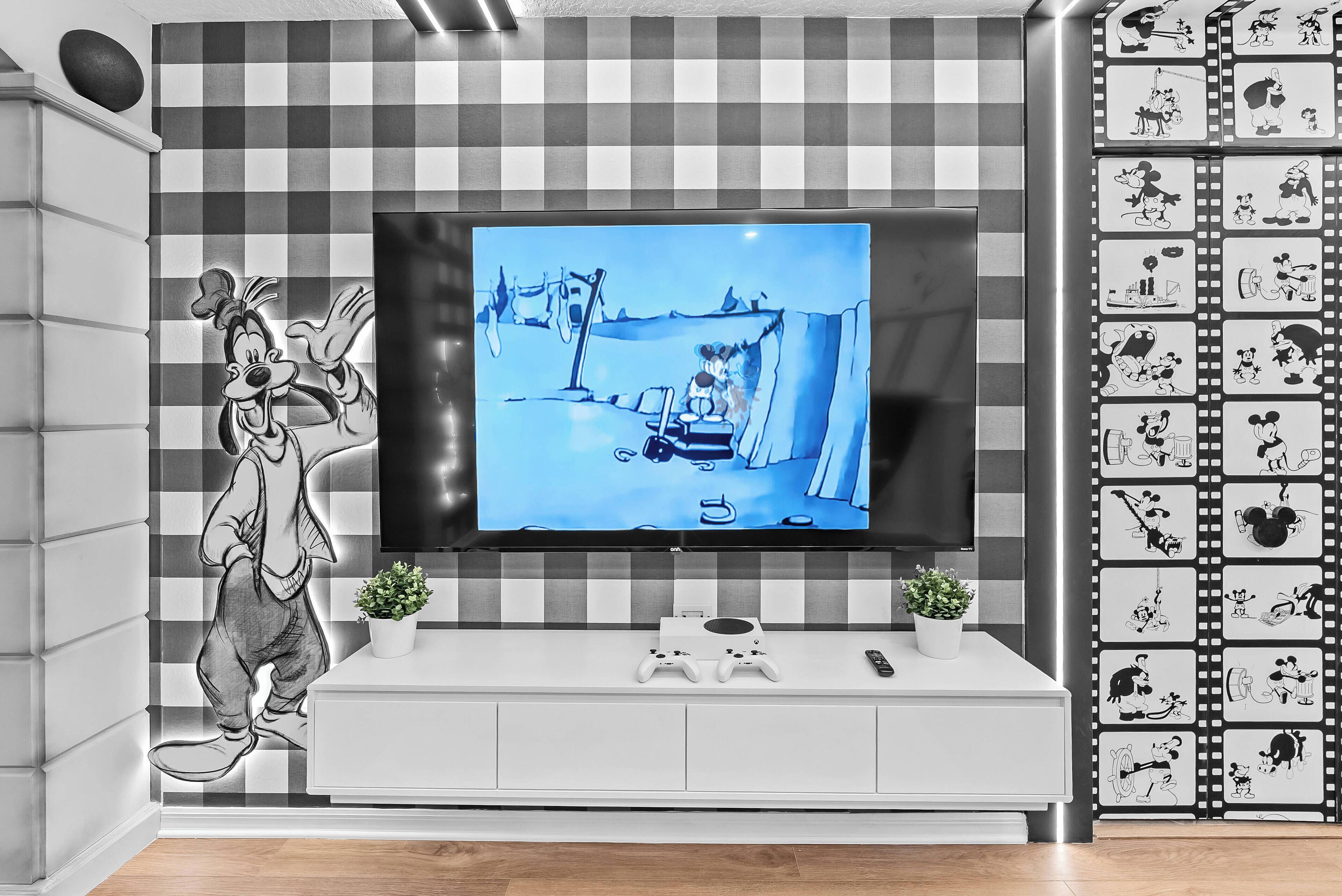 A modern living room with a large flat-screen TV mounted on a wall, a white entertainment unit below, and a Mickey Mouse mural on the left.