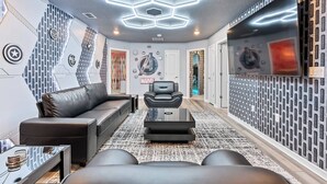 A modern living room with a black leather sofa, a glass coffee table, and a black massage chair. The room features a geometric patterned wall, a large TV, and a ceiling with hexagonal lighting. - 10BR house at Windsor Cay resort, clermont (Clermont)
