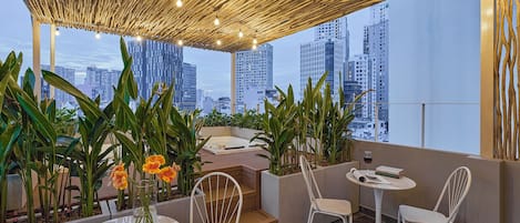 Terraza en la azotea con vistas a edificios de la ciudad, con techo de madera, plantas en macetas y sillas blancas.