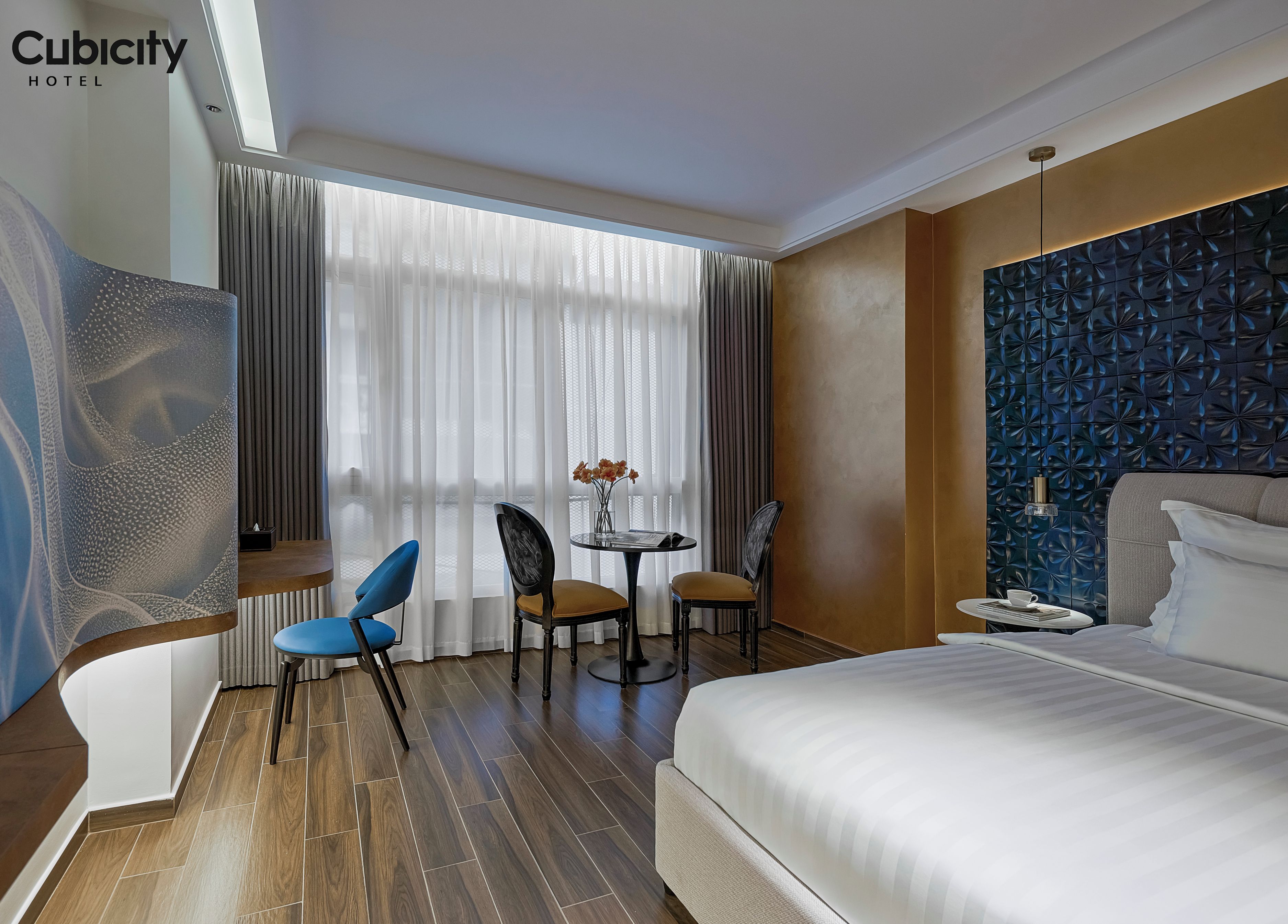 A hotel room with a bed, a desk, two chairs, a small table with a vase, and a window with curtains.