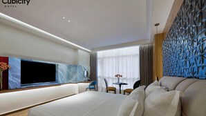 A modern hotel room with a large bed, a flat-screen TV, a small table with chairs, and a decorative wall panel.