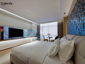 A modern hotel room with a large bed, a flat-screen TV, a small table with chairs, and a decorative wall panel.