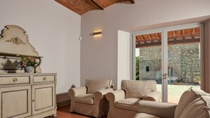 A room with a stone wall, a white cabinet with drawers, beige armchairs, and a large sliding glass door leading to an outdoor area with a stone building.