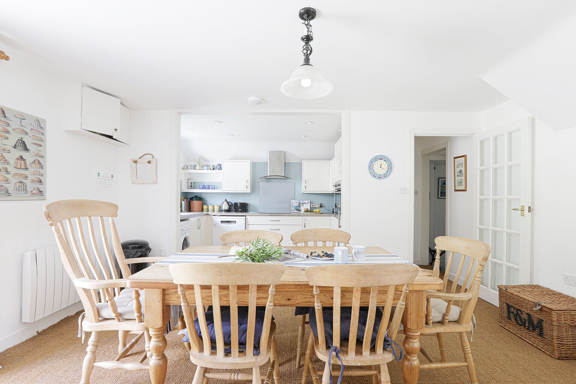 A dining area with a wooden table and chairs, a hanging light fixture, and a kitchen in the background.