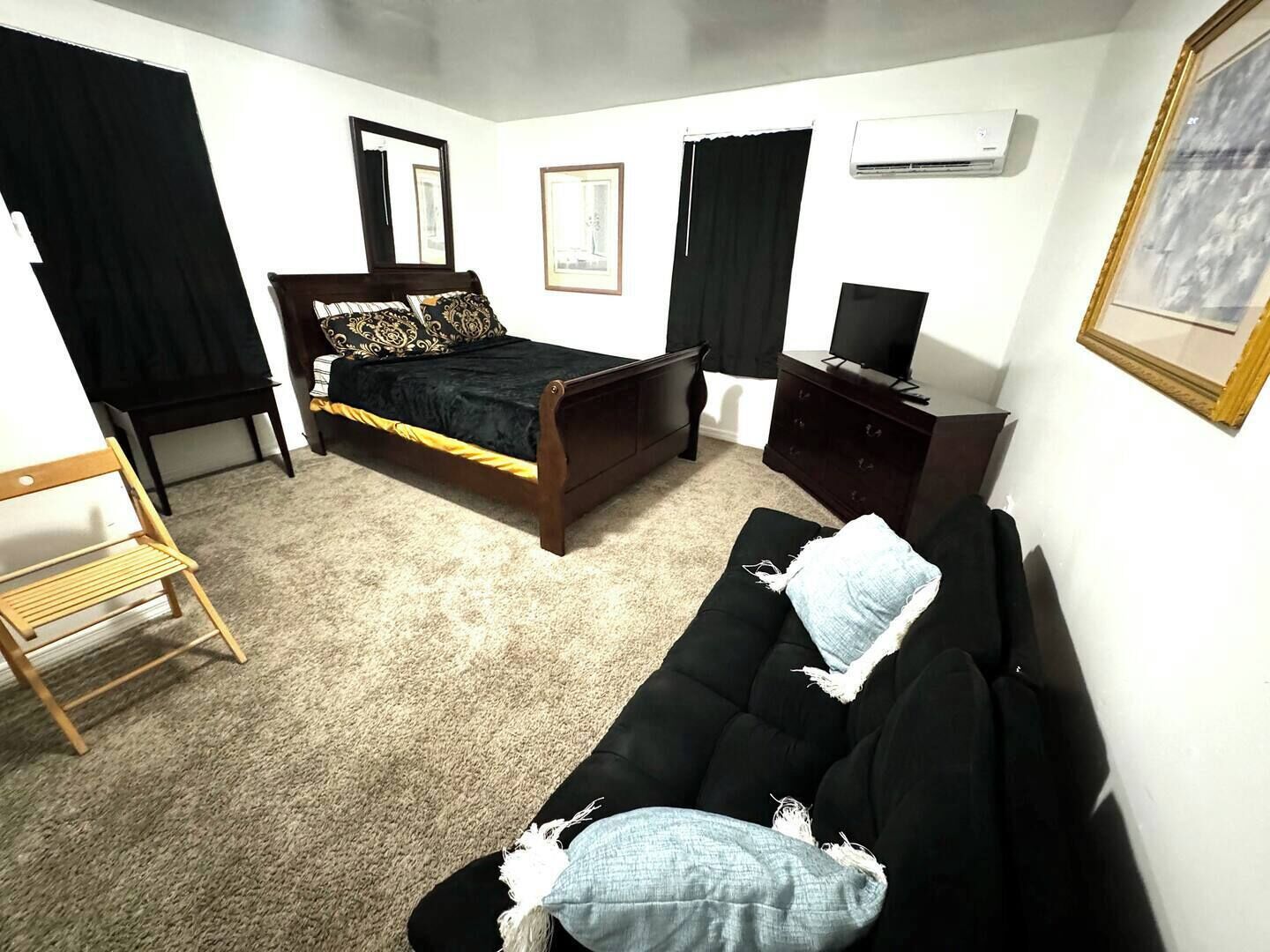 Una camera da letto con un letto, un divano nero, una sedia di legno, una cassettiera e una televisione.