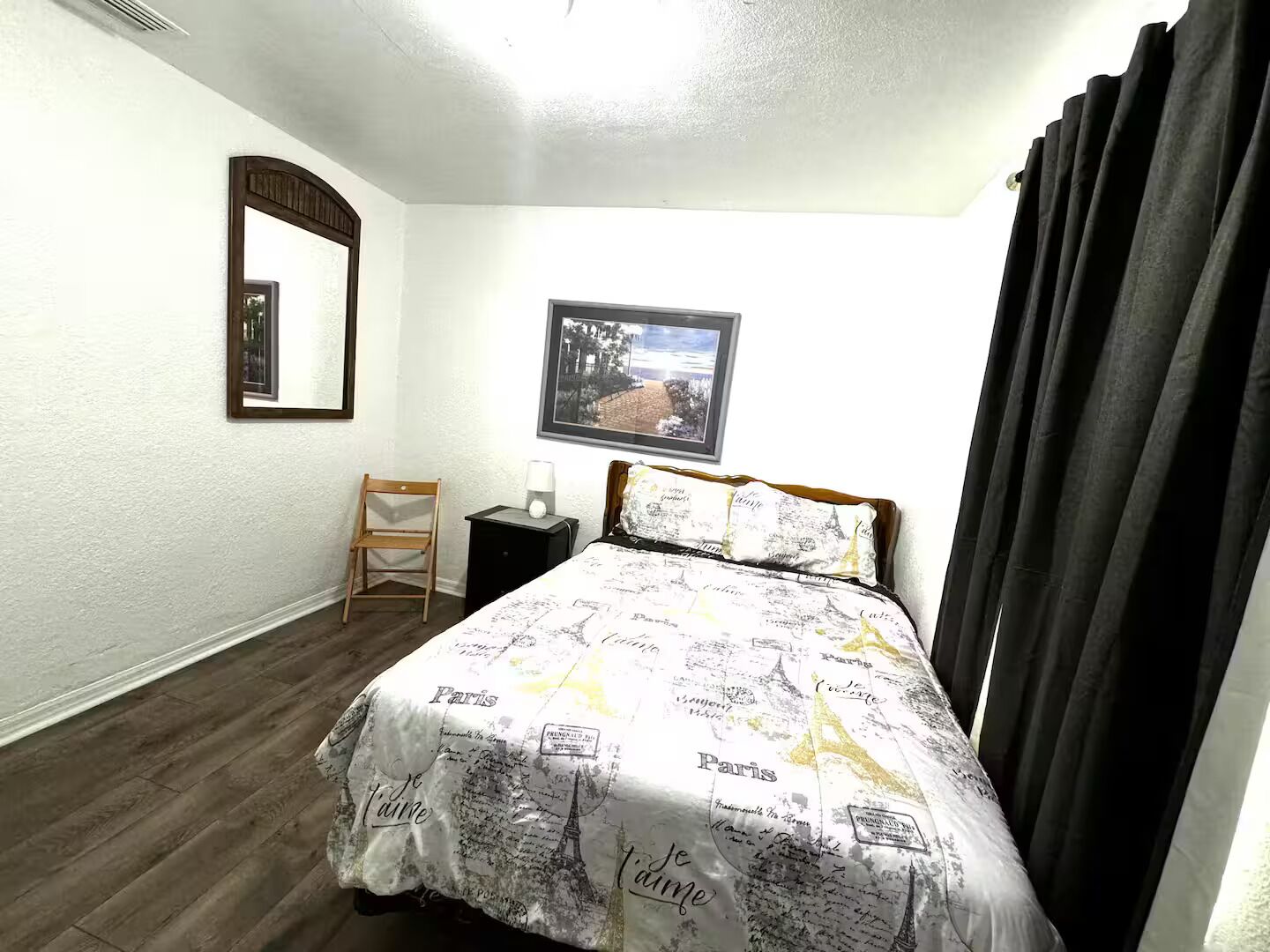 Ein Schlafzimmer mit Bett, Stuhl, Nachttisch, Spiegel und einem gerahmten Bild an der Wand.