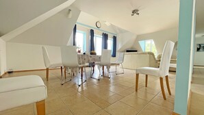Amplia habitación en el ático con comedor, muebles blancos y un gran ventanal con cortinas.