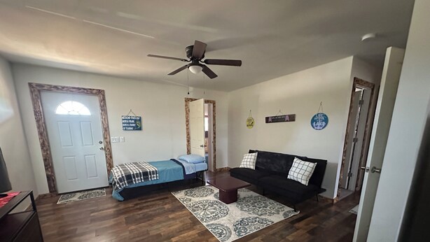 Una habitación con una cama, un sofá negro, una mesa de centro, un ventilador de techo y una alfombra estampada.