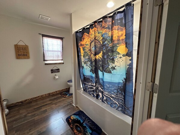 A hallway with a wooden floor, a window with a blind, a toilet, and a shower curtain with a tree design.
