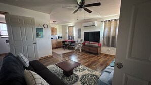 Sala de estar con sofá, mesa de centro y alfombra estampada. Hay un comedor con mesa y sillas, además de una cocina con armarios y electrodomésticos. Se observa un ventilador de techo.