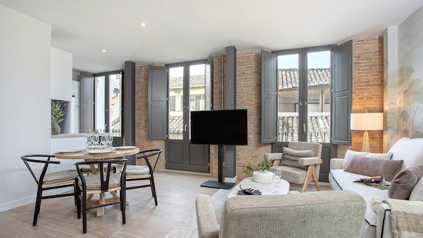 A modern living room with a dining area, a flat-screen TV, and large windows with shutters.