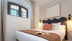 Ein ordentlich bezogenes Bett mit brauner Decke, weißer Bettwäsche und einem Buch darauf. Ein Fenster mit Vorhängen, ein an der Wand befestigtes Kunstwerk und eine Nachttischlampe sind ebenfalls zu sehen.