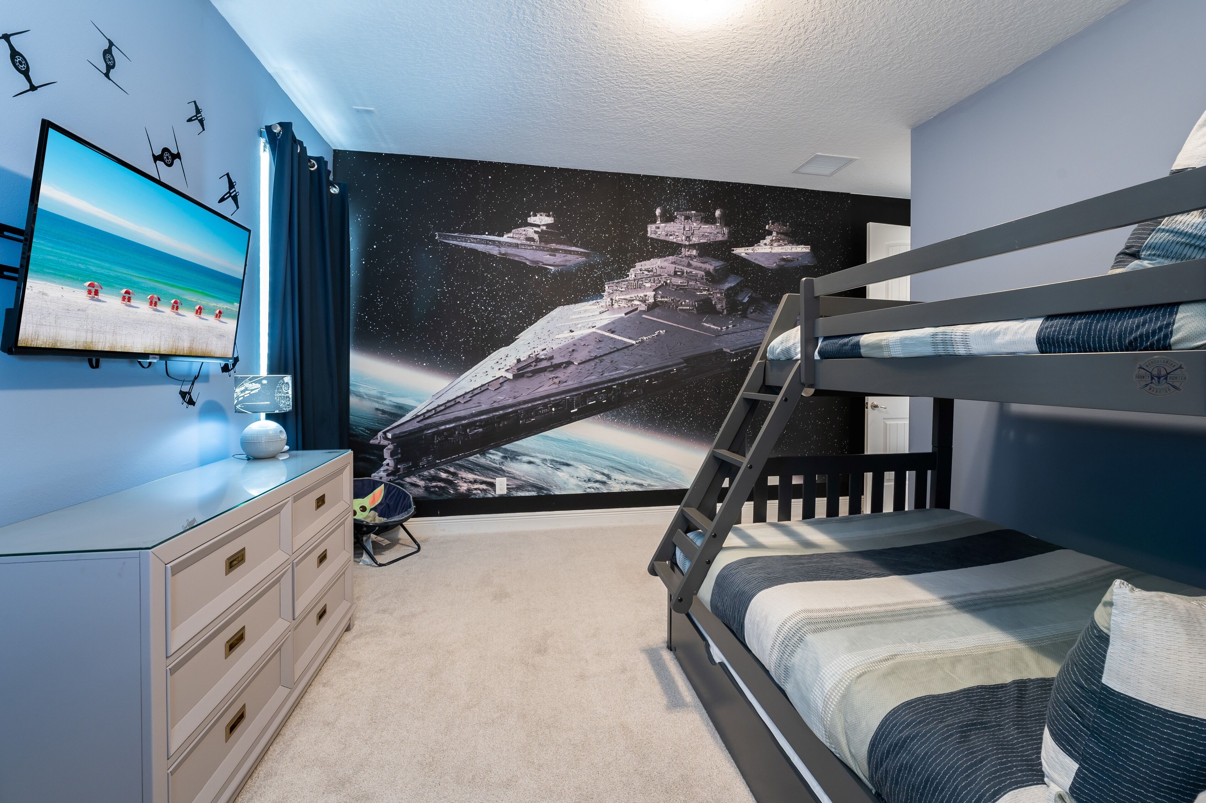 Un dormitorio con una cama litera, un televisor montado en la pared, un aparador y un mural con motivos espaciales.