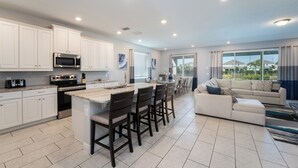 A modern kitchen with white cabinets, a central island with bar stools, a built-in microwave, and a stove. There is a dining area with a white sofa and a large window providing a view of the outside. - Discover Your Inner Child  Encore Villa (Kissimmee)