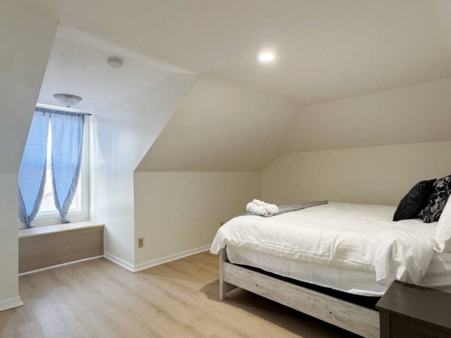 A bedroom with a sloped ceiling, a bed with white bedding, a window with blue curtains, and a small bedside table.
