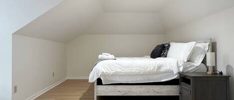 A bedroom with a sloped ceiling, a bed with white bedding, a bedside table with a lamp, and a small cabinet.