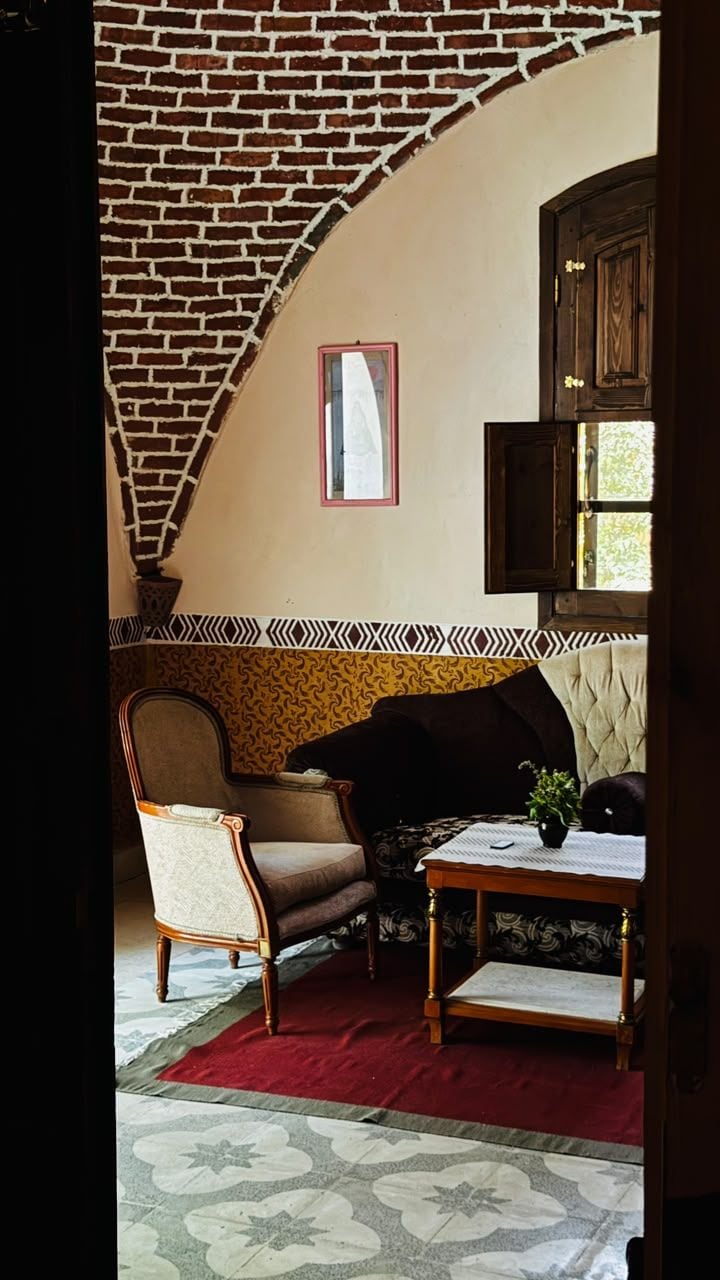 Een kamer met een bakstenen boog, een behang met patroon, een bank, een stoel en een kleine tafel met een plant.