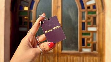Una mano sostiene una llave de acceso con la inscripción "Habu Villa", frente a una puerta de madera con vidrieras de colores.