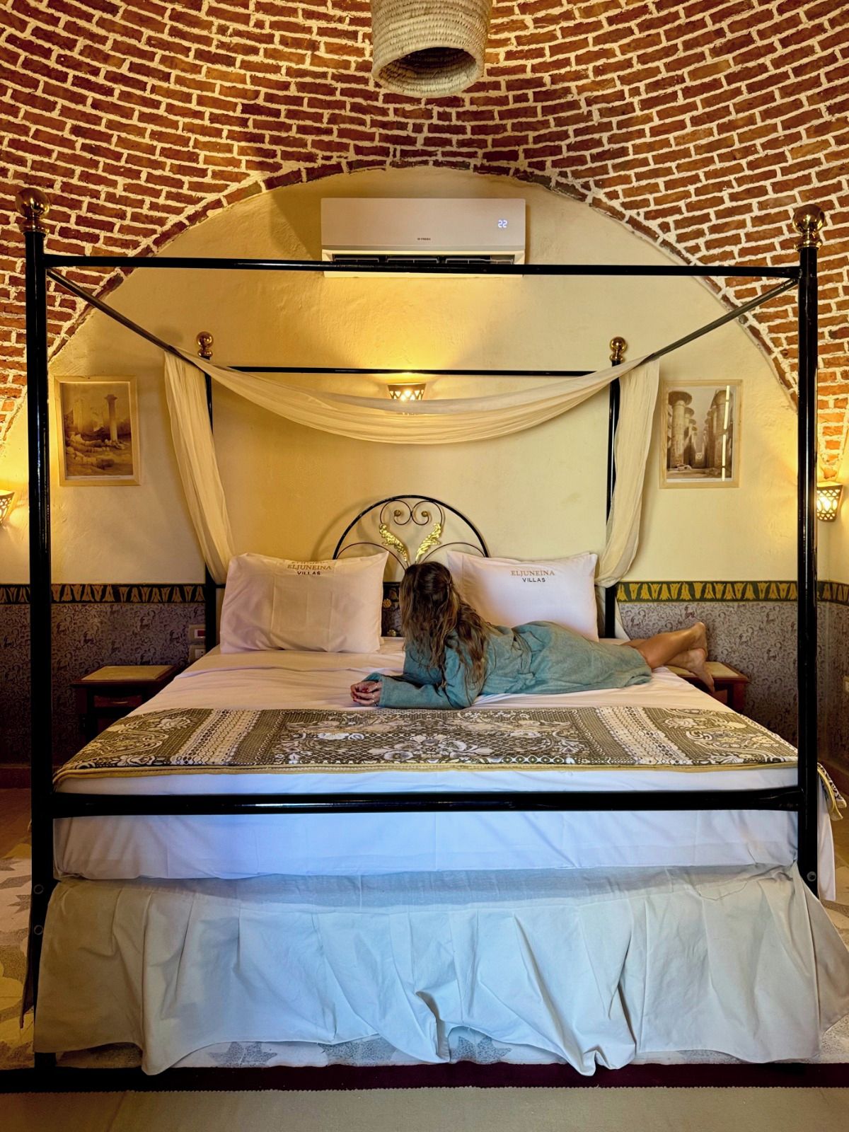 Una cama con dosel de madera con un edredón y almohadas blancos, estructura metálica negra y pared de ladrillo al fondo.