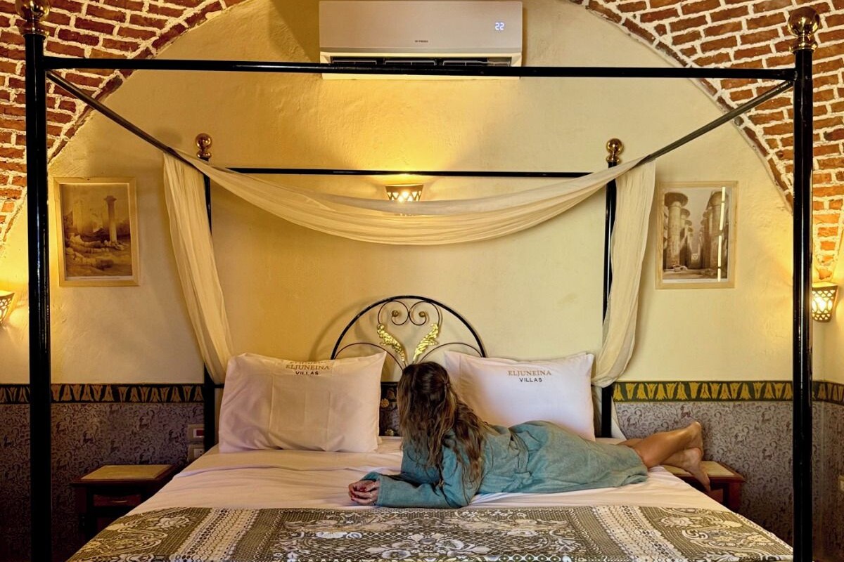 Een hemelbed met een witte spreideken en kussens, een zwart metalen frame en een achtergrond van een bakstenen muur.